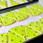 Bright green Christmas tree sugar cookies lined up on a baking tray, with different colored sugar pearls as ornaments.